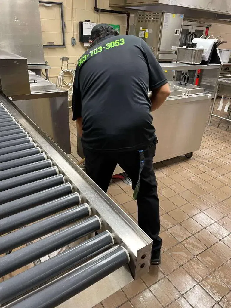 Man in black uniform working on kitchen equipment near a conveyor belt.