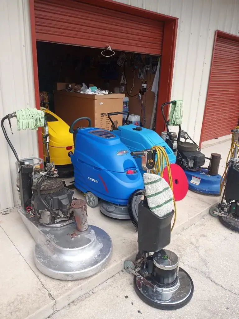 Floor cleaning machines of various sizes, colors, and types are stored near an open garage door.