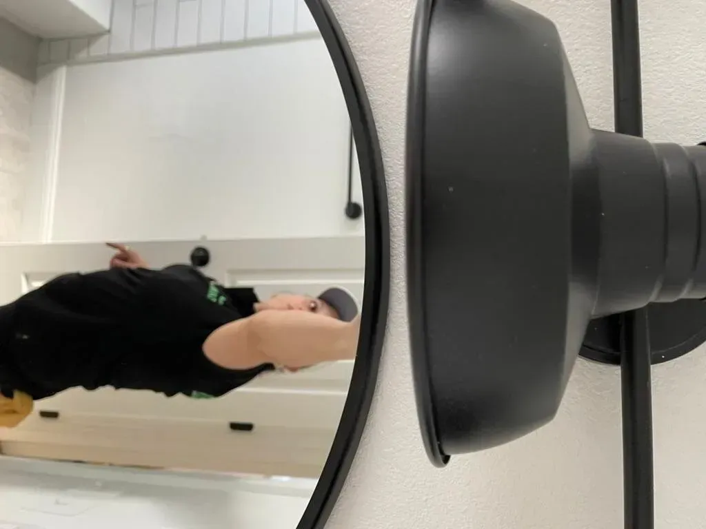 A person in black clothing is reflected in a round mirror, next to a black light fixture, on a white wall.