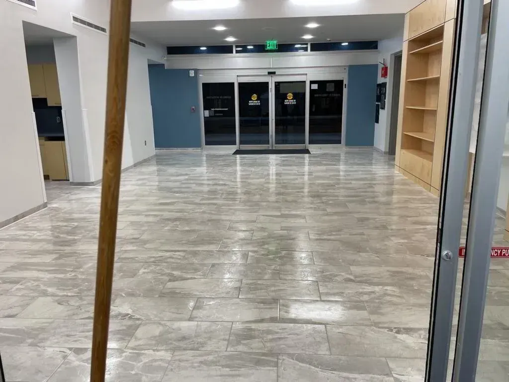Empty, tiled lobby with automatic doors, blue wall, and partially built shelving. A broom leans in the foreground.