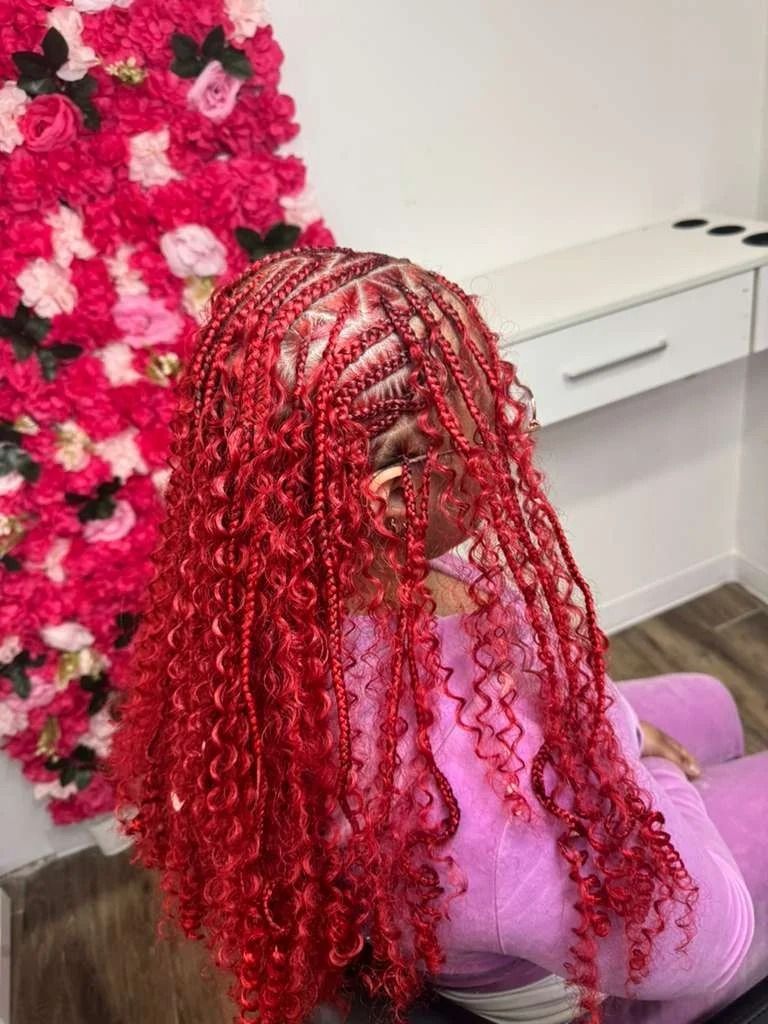 Person with long red curly braids seated beside a pink floral wall in a salon