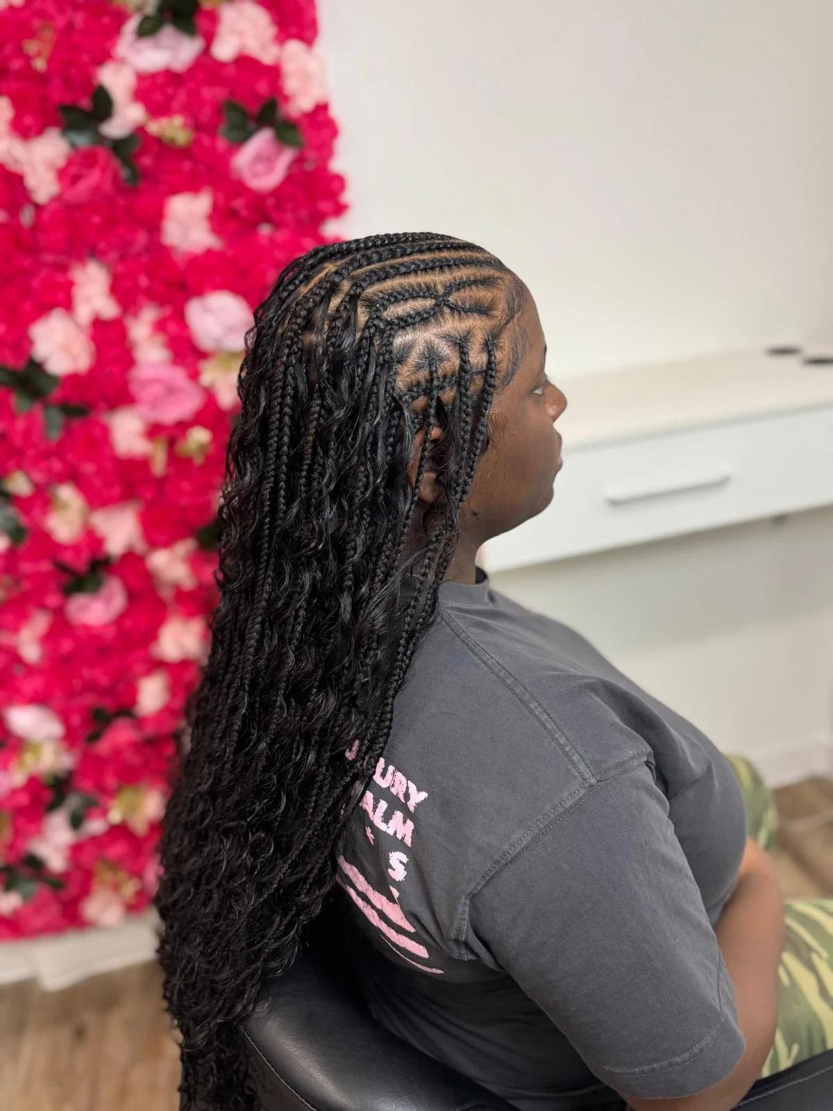 Woman with long braided hair seated indoors, side view, against a white wall and pink floral backdrop.