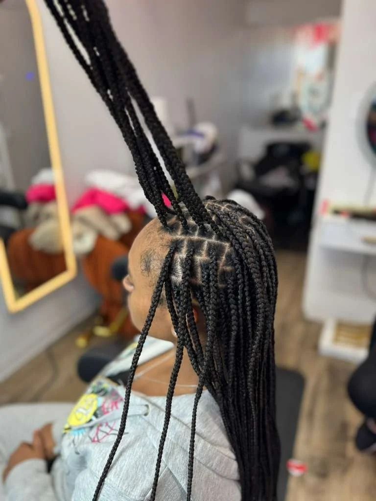 Side view of a person with long braided hair in a salon chair, with mirrors and styling tools in the background.