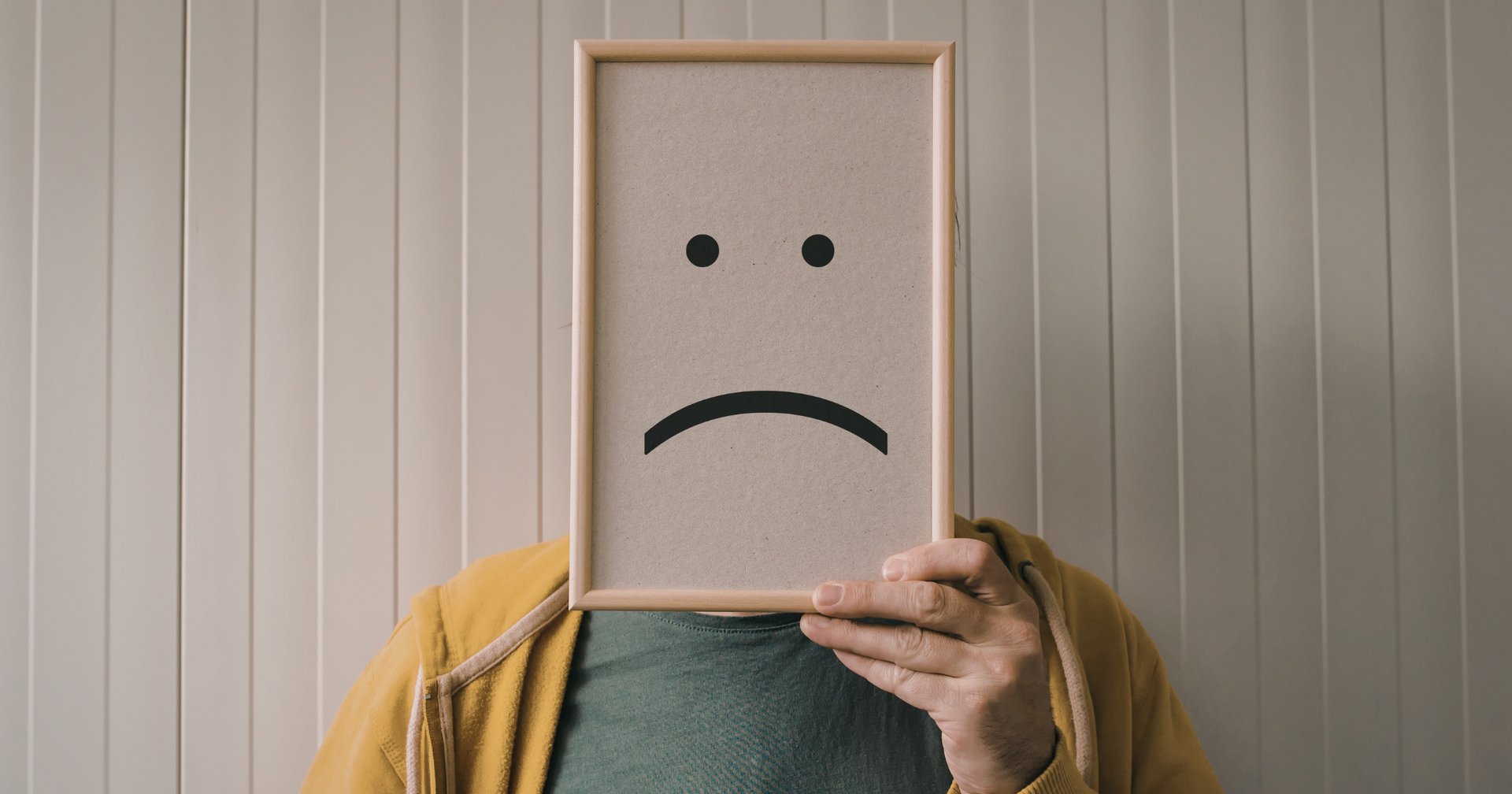 Person holding a frame with a sad face drawn on it; yellow jacket, green shirt.
