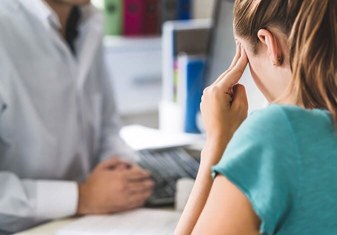 Una mujer está sentada en un escritorio con la cabeza entre las manos mientras habla con un médico.