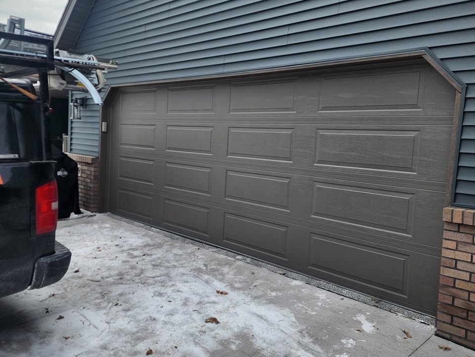 Brown garage door on a house with snowy driveway and a vehicle parked beside it.