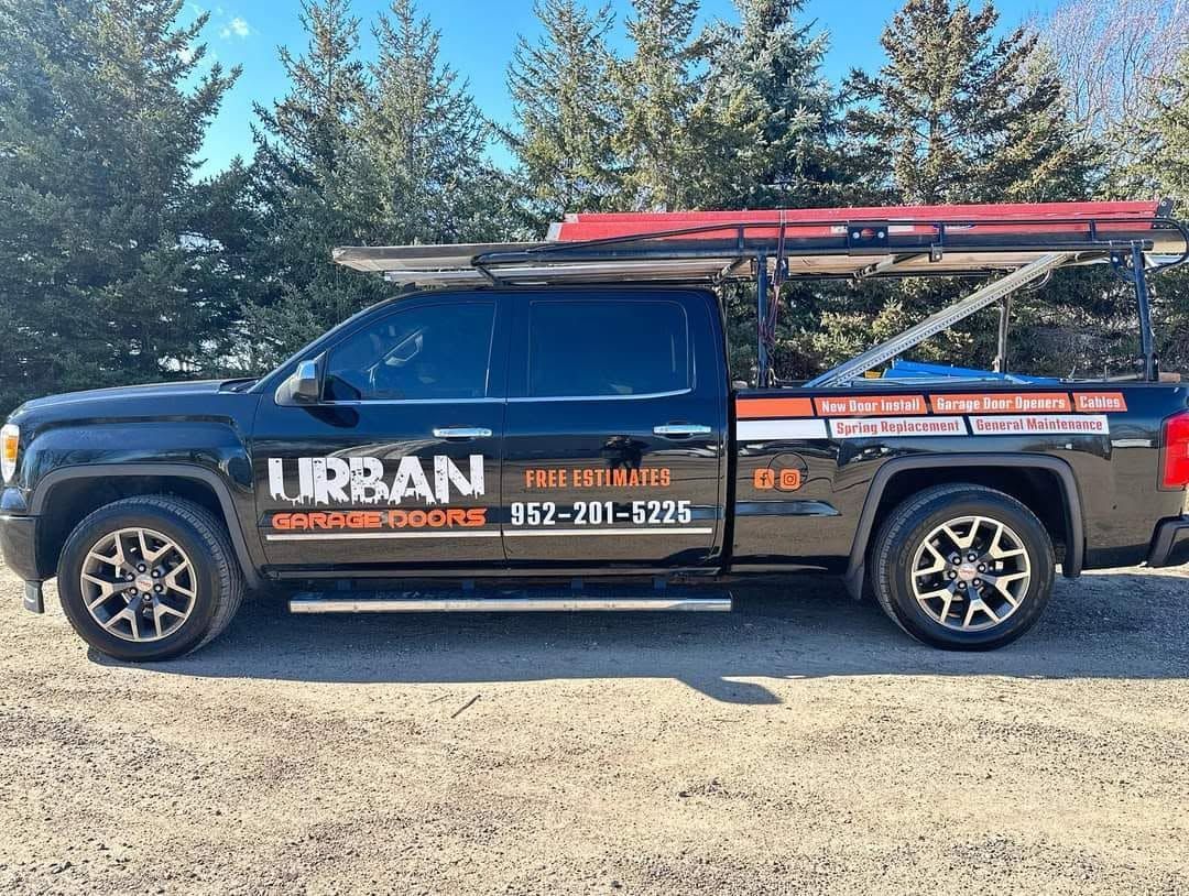 Black URBAN service pickup truck with ladders on top, parked outdoors on a gravel lot.