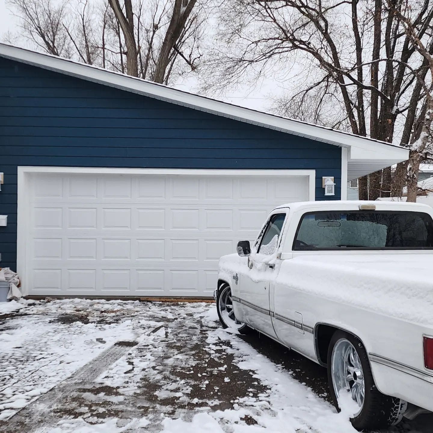 White pickup truck parked in snowy driveway beside a blue house with a white garage door
