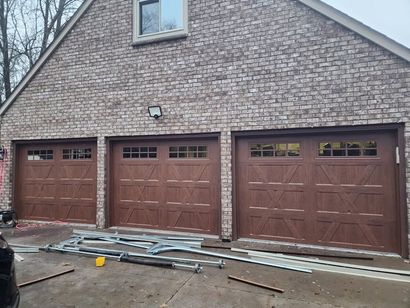 Three brown garage doors on a brick house exterior, with construction materials on the ground.