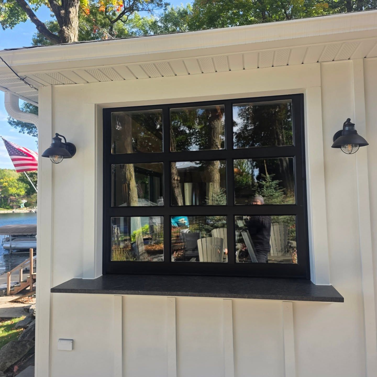 Black-framed window in a white house exterior with dark wall lanterns and a lake reflection in the glass