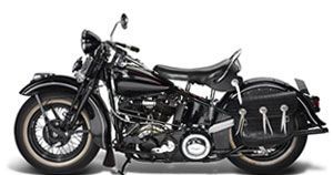 Black vintage Harley-Davidson motorcycle with white-walled tires, leather saddlebags, and a dark backdrop.