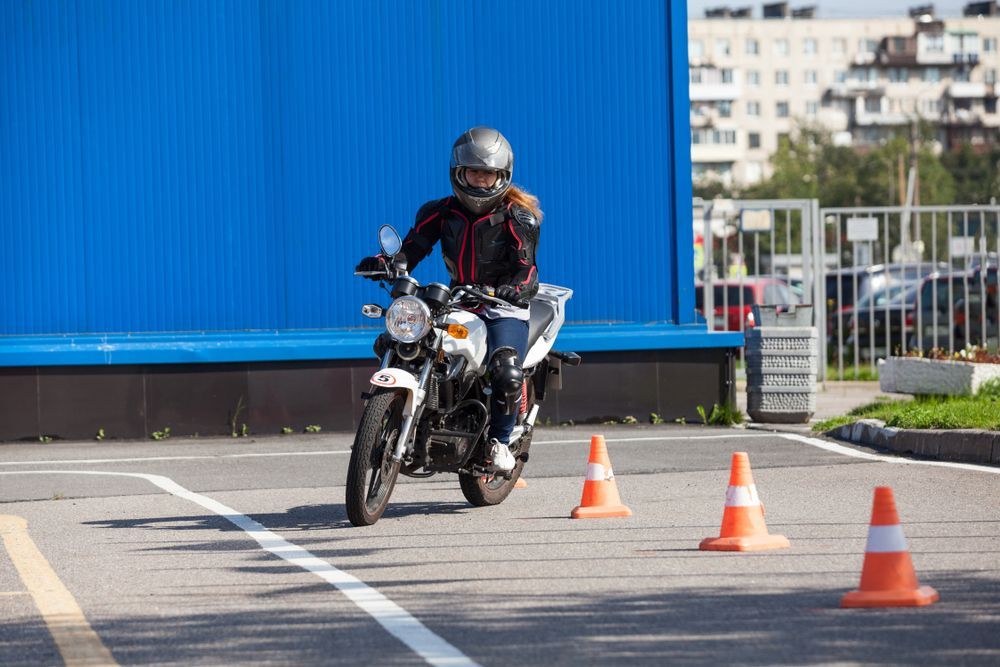 Person on motorcycle maneuvers through cones in a parking lot, wearing helmet and protective gear.