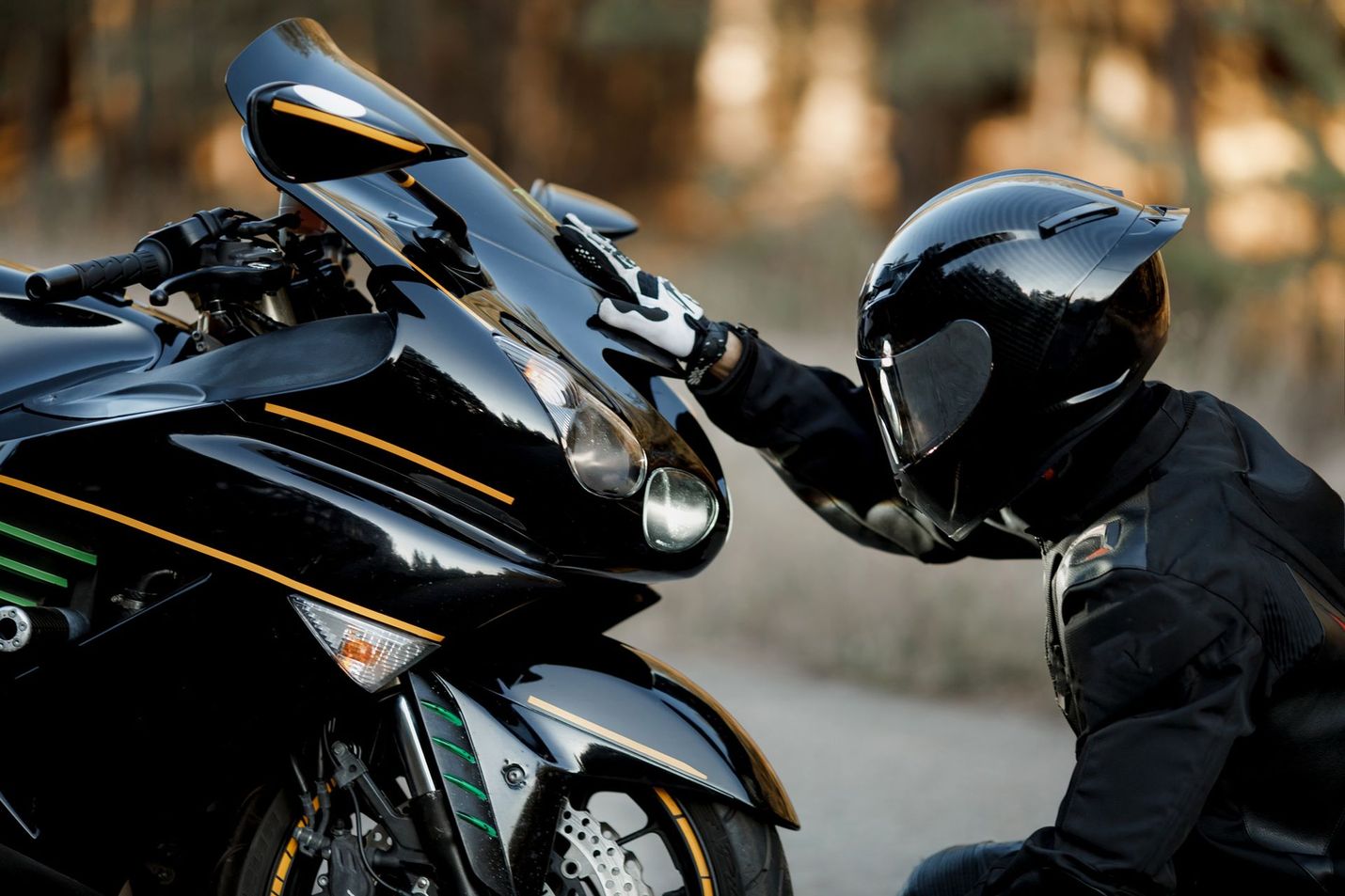 Person in black helmet and gear touching a black motorcycle outdoors, sunlight.