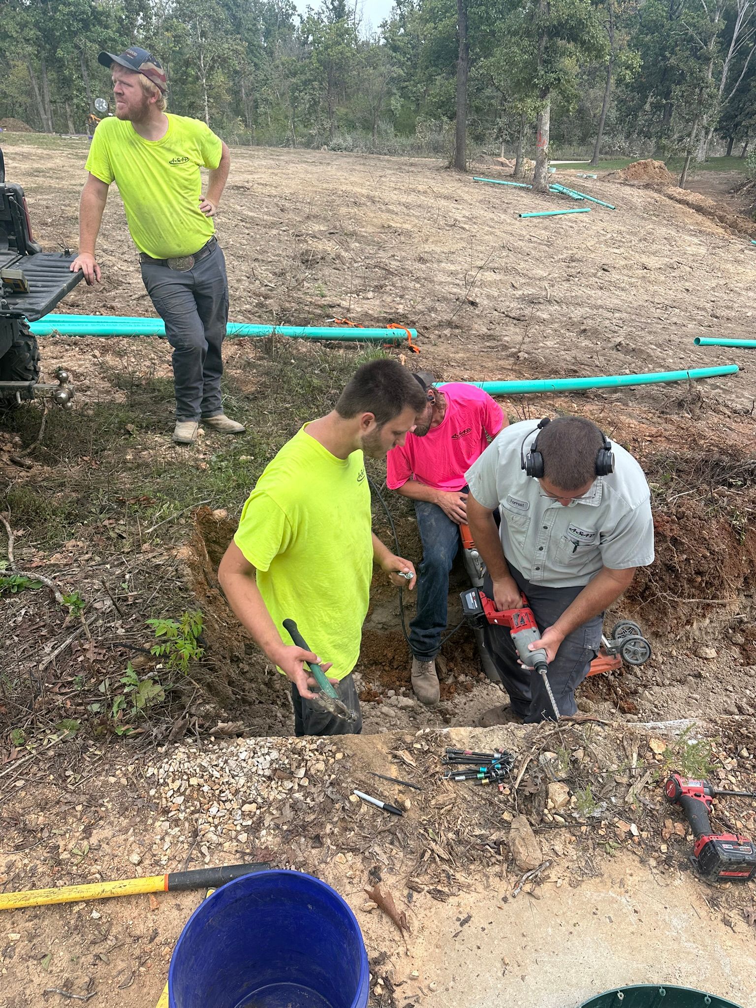Two workers install white pipes with purple caps in a muddy outdoor trench.