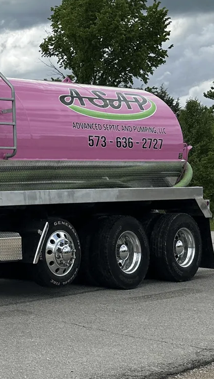 Pink and green tanker truck viewed from the rear on a gravel road with trees in the background