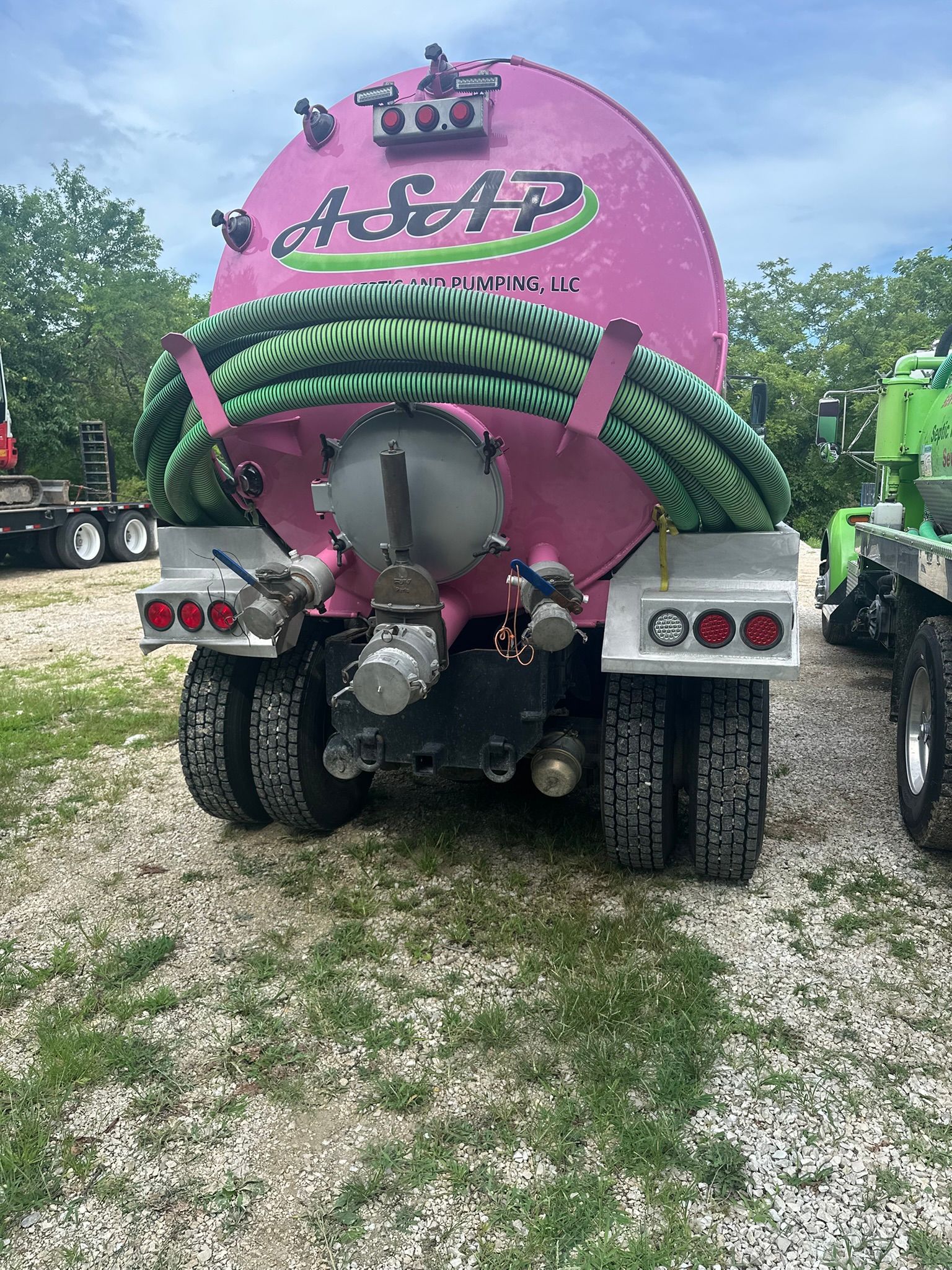 Pink and green tanker truck viewed from the rear on a gravel road with trees in the background