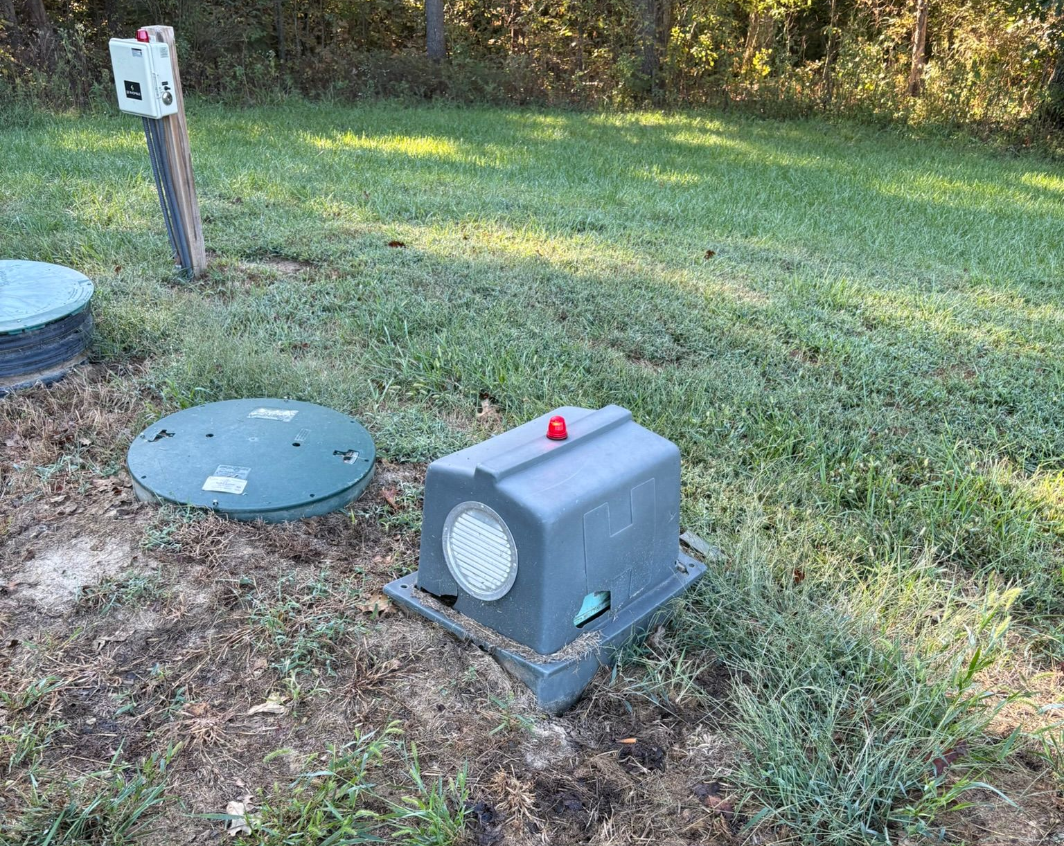 Gray utility box with red marker on a grassy lawn beside a circular green cover and a post