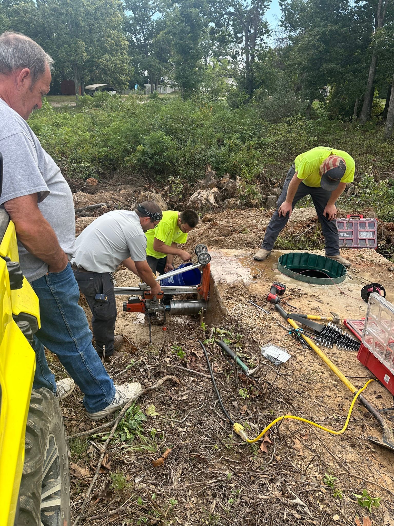 Workers in neon shirts dig around a ground opening in a wooded outdoor area with tools and cables.