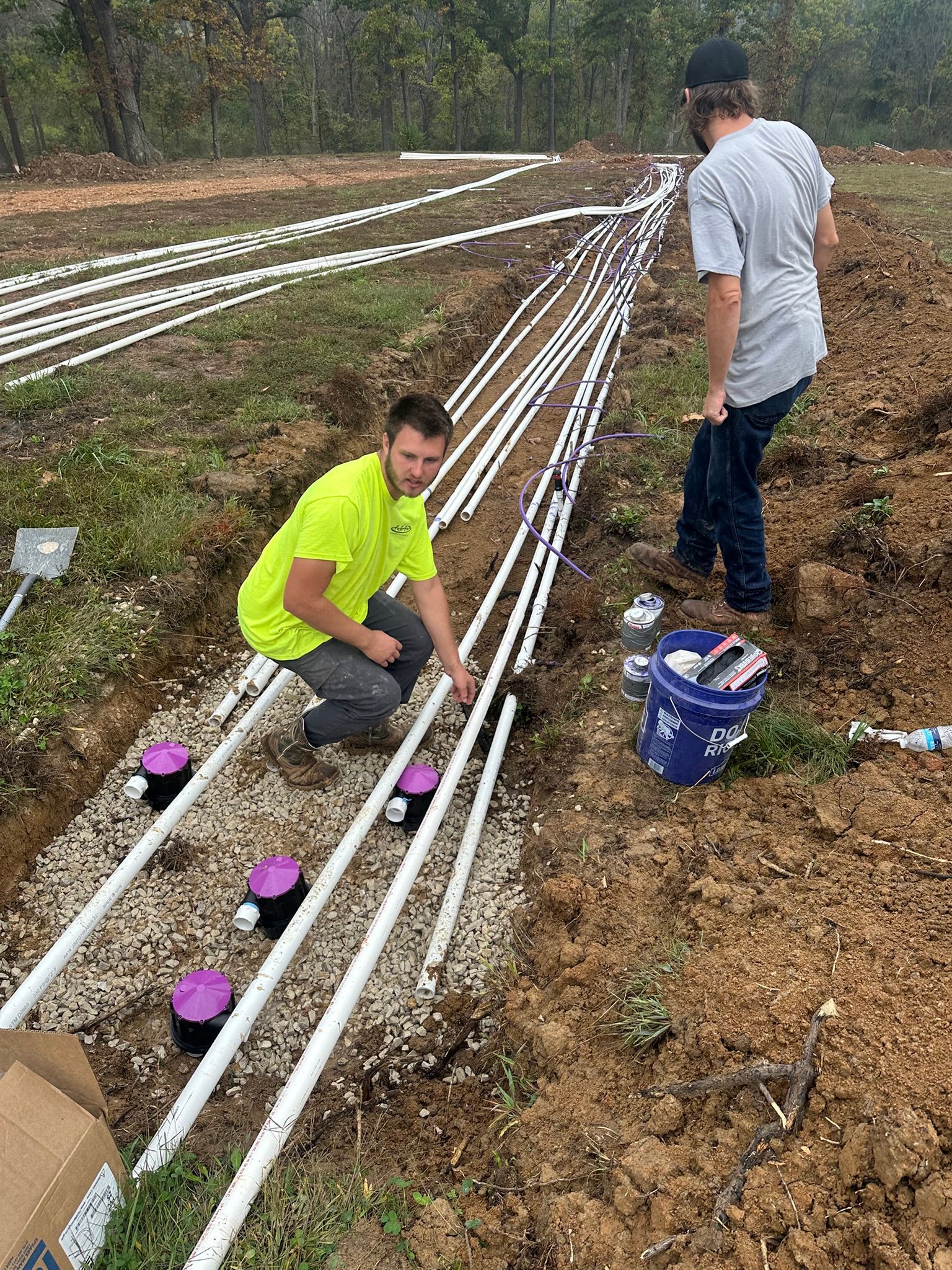 Two people installing white pipes with purple caps along a dirt trench outdoors
