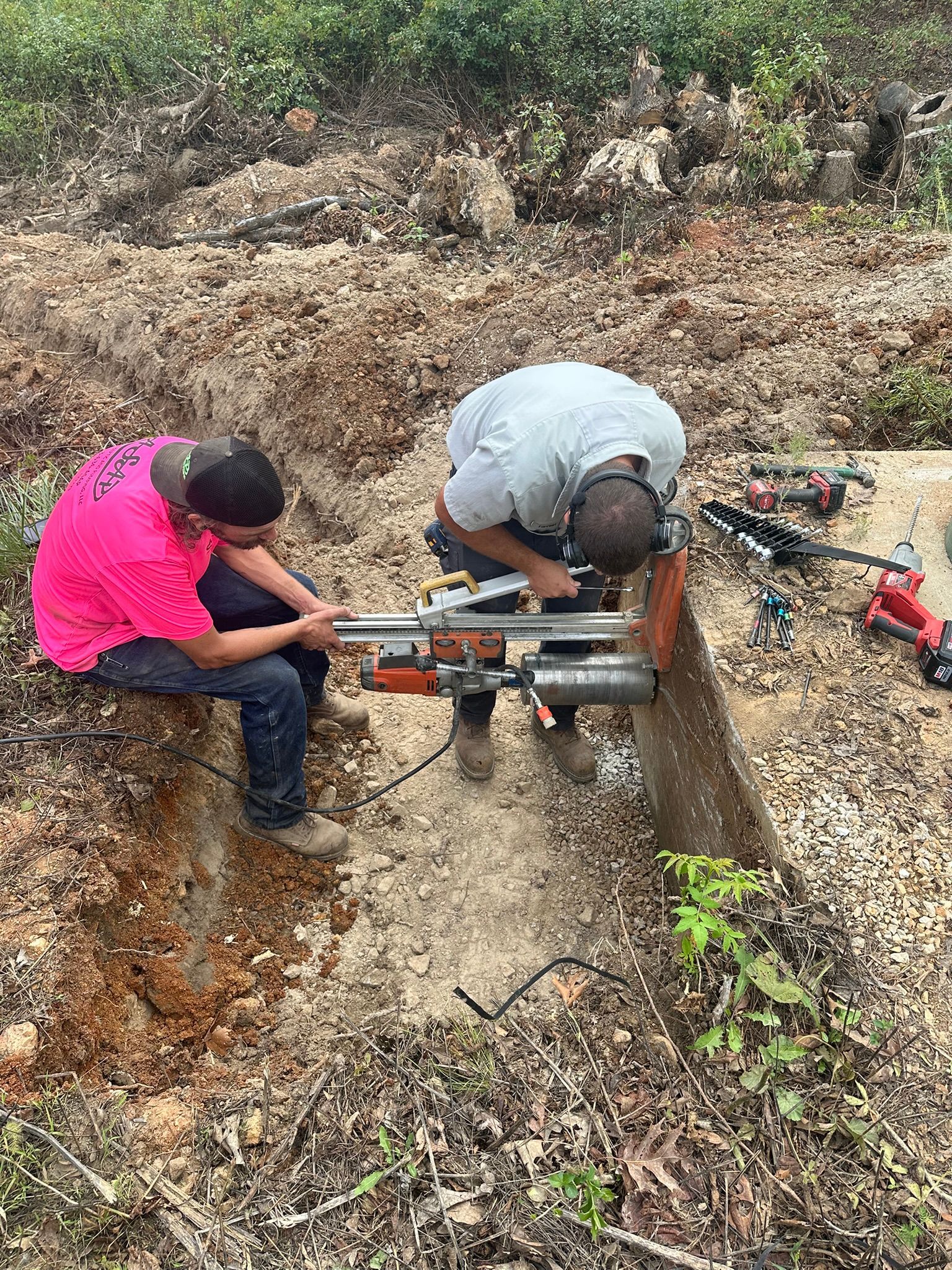Two workers crouch over a trench, using a tool to cut or repair a pipe in a dirt ditch.