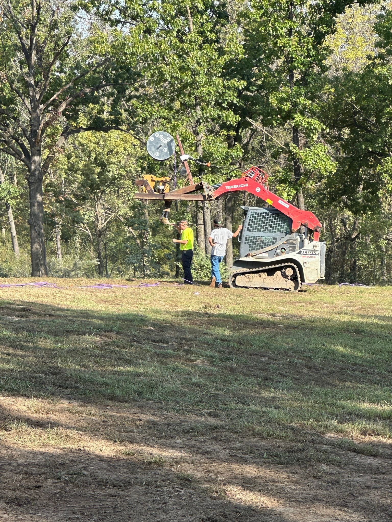 Excavator lifting a large rust-colored metal tank at a dirt construction site, with trees in the background
