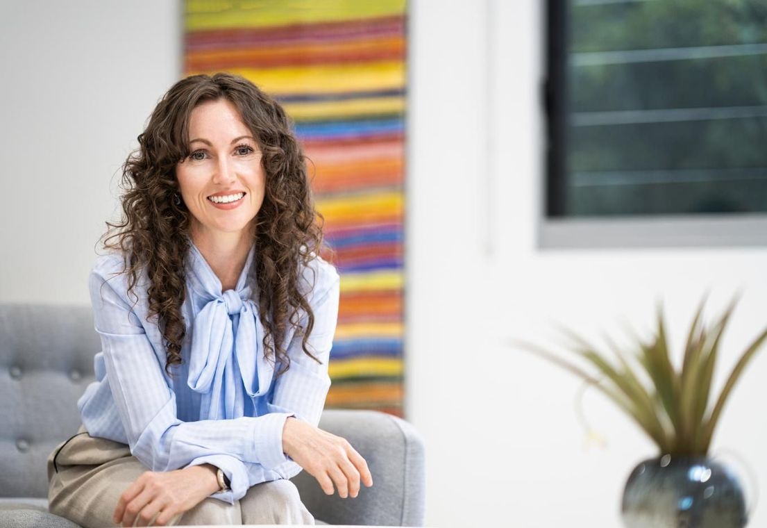Woman smiling on a couch in a bright room with a colorful wall hanging and potted plant.