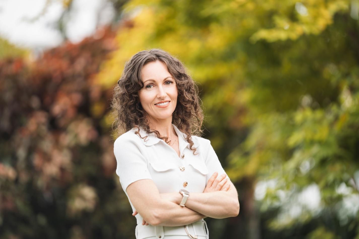 Smiling woman with curly hair standing outdoors with arms crossed, wearing a white blouse, blurred greenery behind her