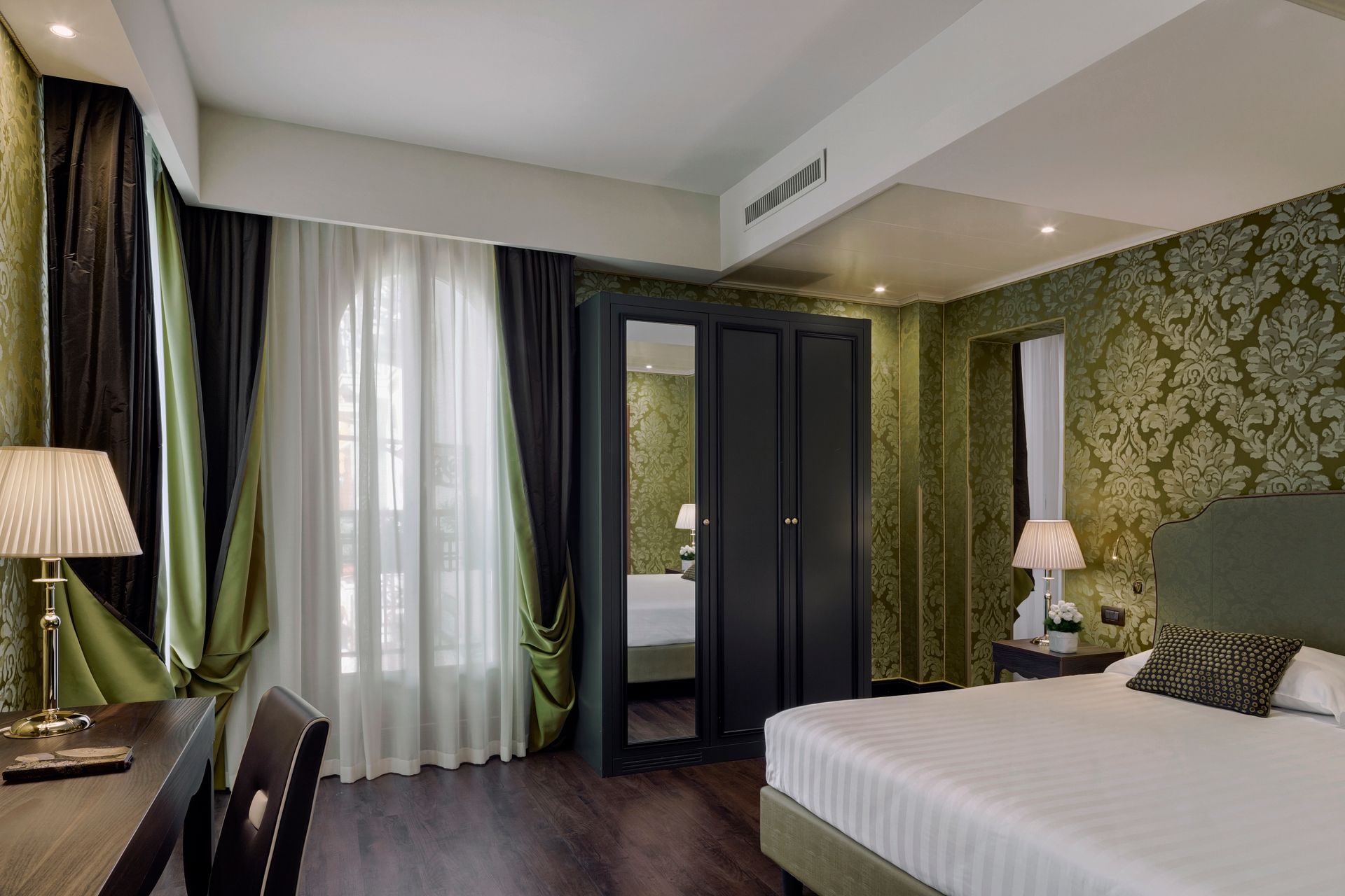 Camera d'albergo moderna con letto, carta da parati a motivi geometrici, tende scure e zona salotto con finestra.