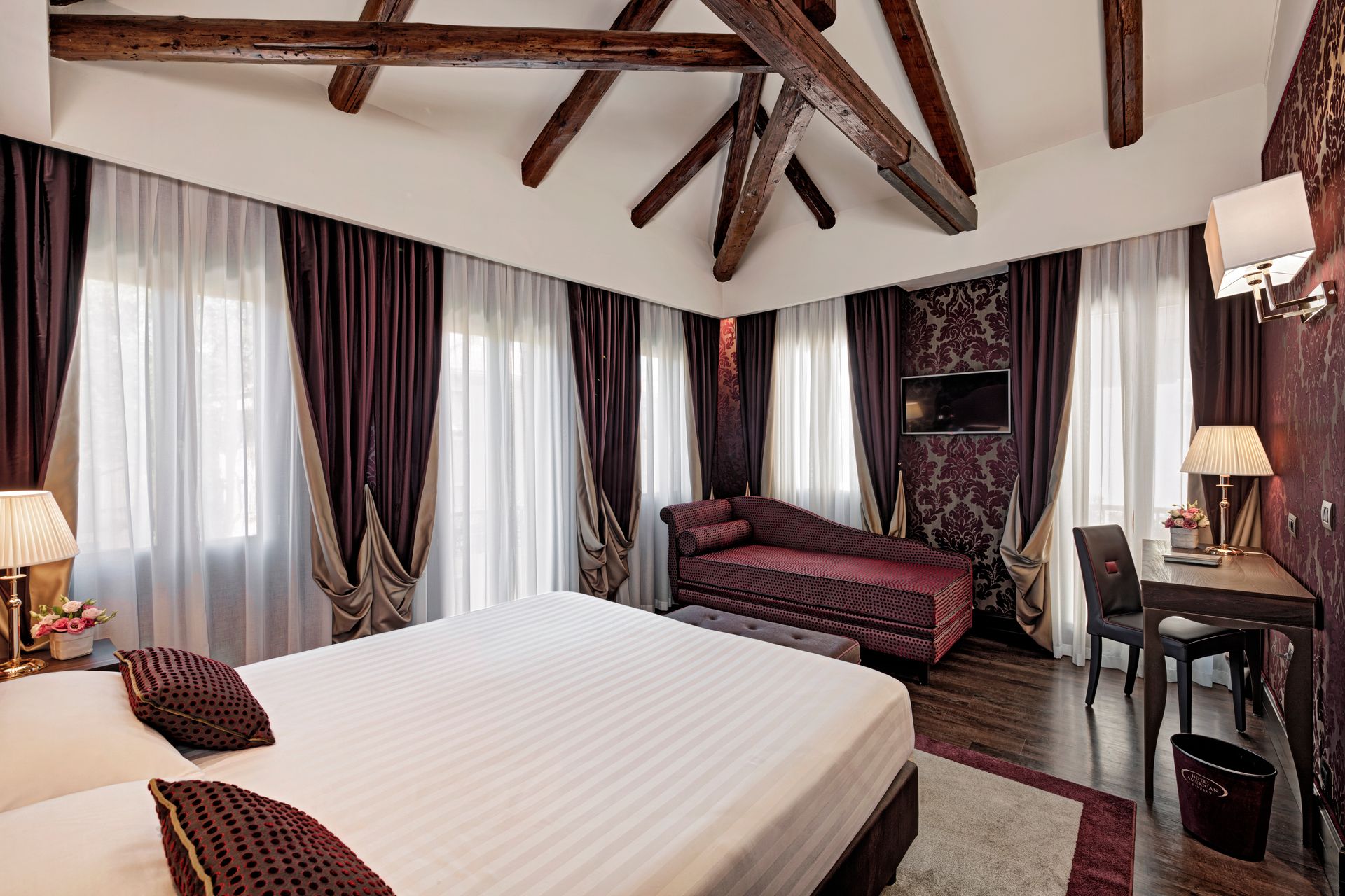 Elegante camera d'albergo con letto bianco, tende scure, travi in ​​legno a vista e un angolo salotto.