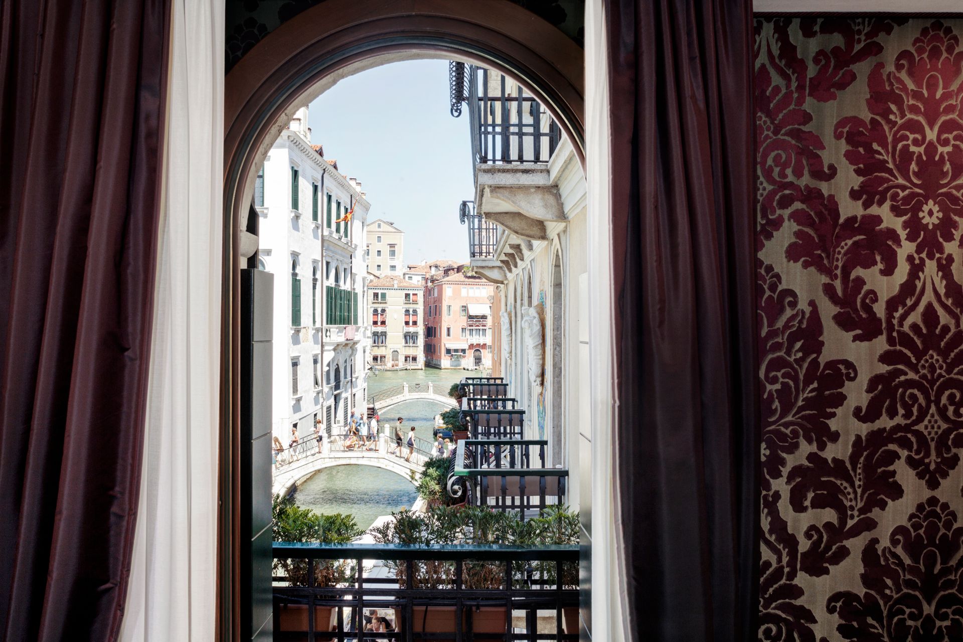 Vista attraverso una finestra ad arco con tende rosse su un canale veneziano e sugli edifici sullo sfondo.