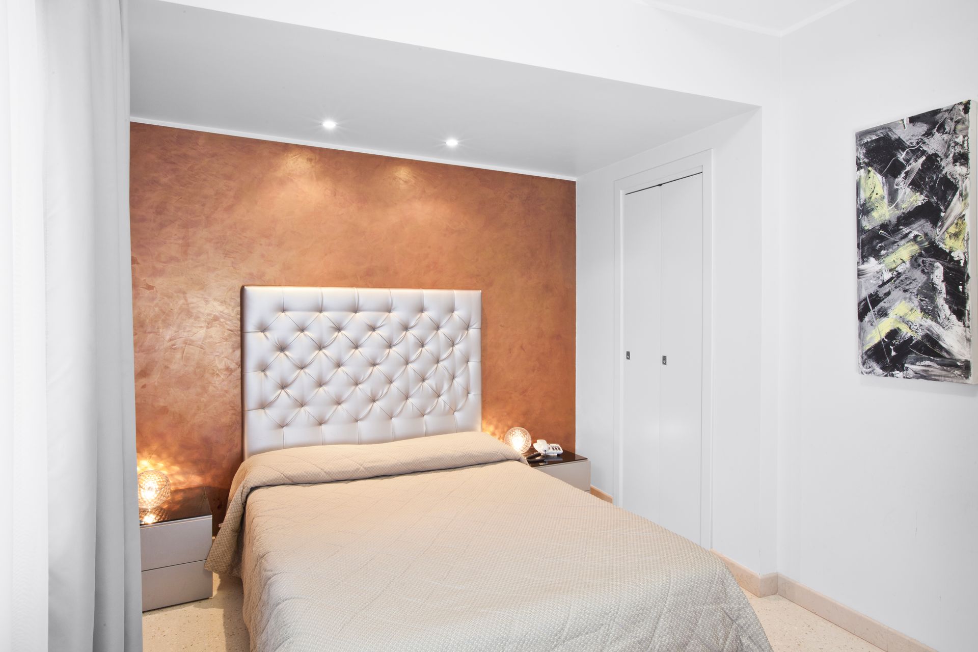 Modernes Schlafzimmer mit Bett, weißem, gestepptem Kopfteil, kupferfarbener Akzentwand und Wandbild.