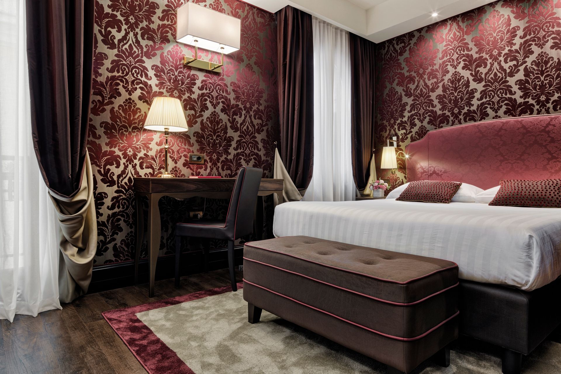 Elegante camera da letto color bordeaux con carta da parati a motivi geometrici, un grande letto, scrivania, tende e panca