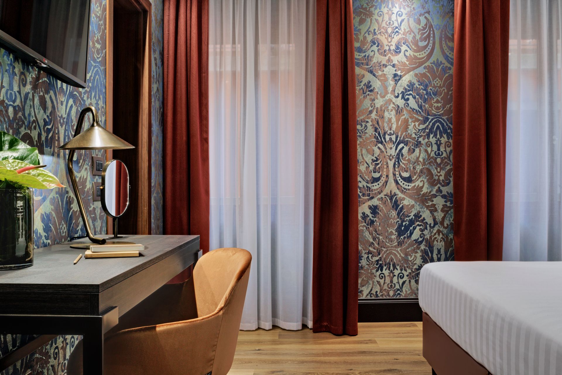 Elegante camera d'albergo con carta da parati a motivi, scrivania e sedia, letto e alta finestra con tende.