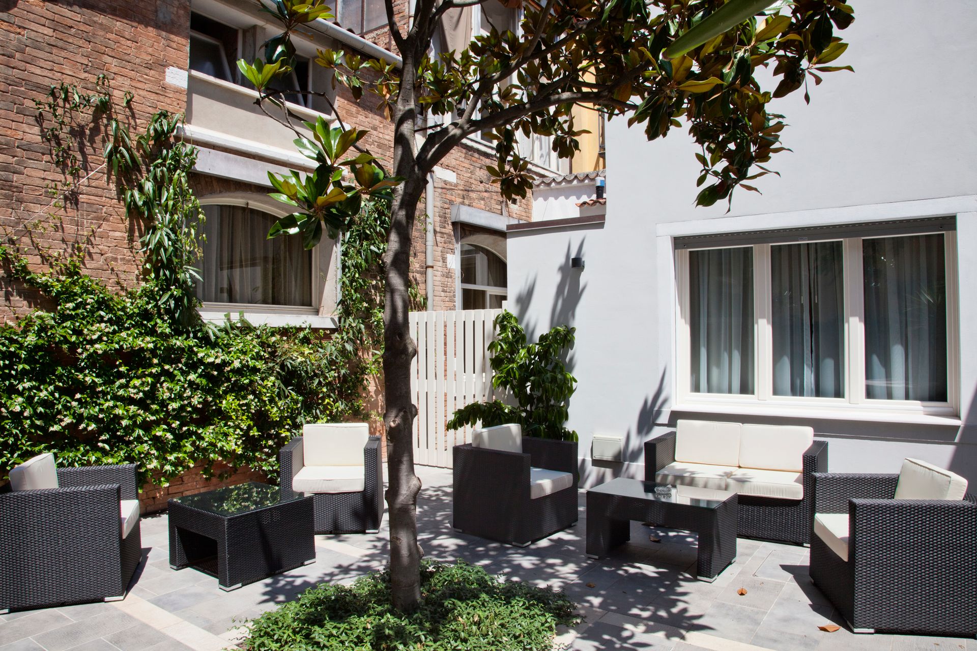 Cortile con patio arredato con sedie e tavoli in vimini, accanto a edifici in mattoni e bianchi, ombreggiato da un albero.