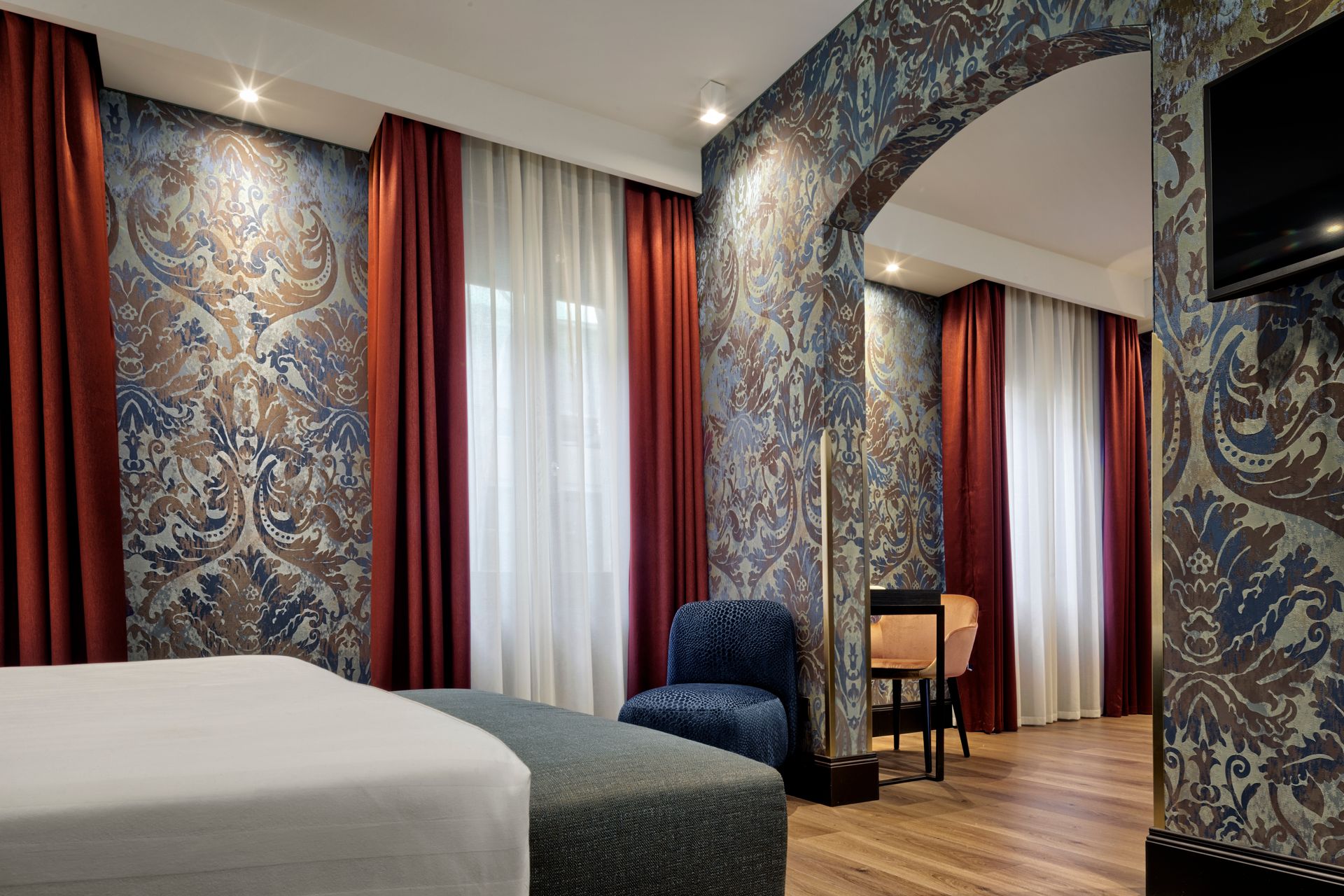 Elegante camera d'albergo con carta da parati decorata blu-rossa, letto, poltrona e tende alle finestre.