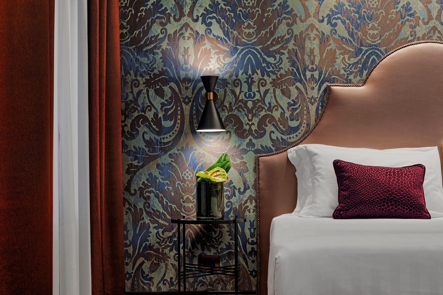 Camera d'albergo con carta da parati a motivi, testiera color crema, lenzuola bianche e applique a muro