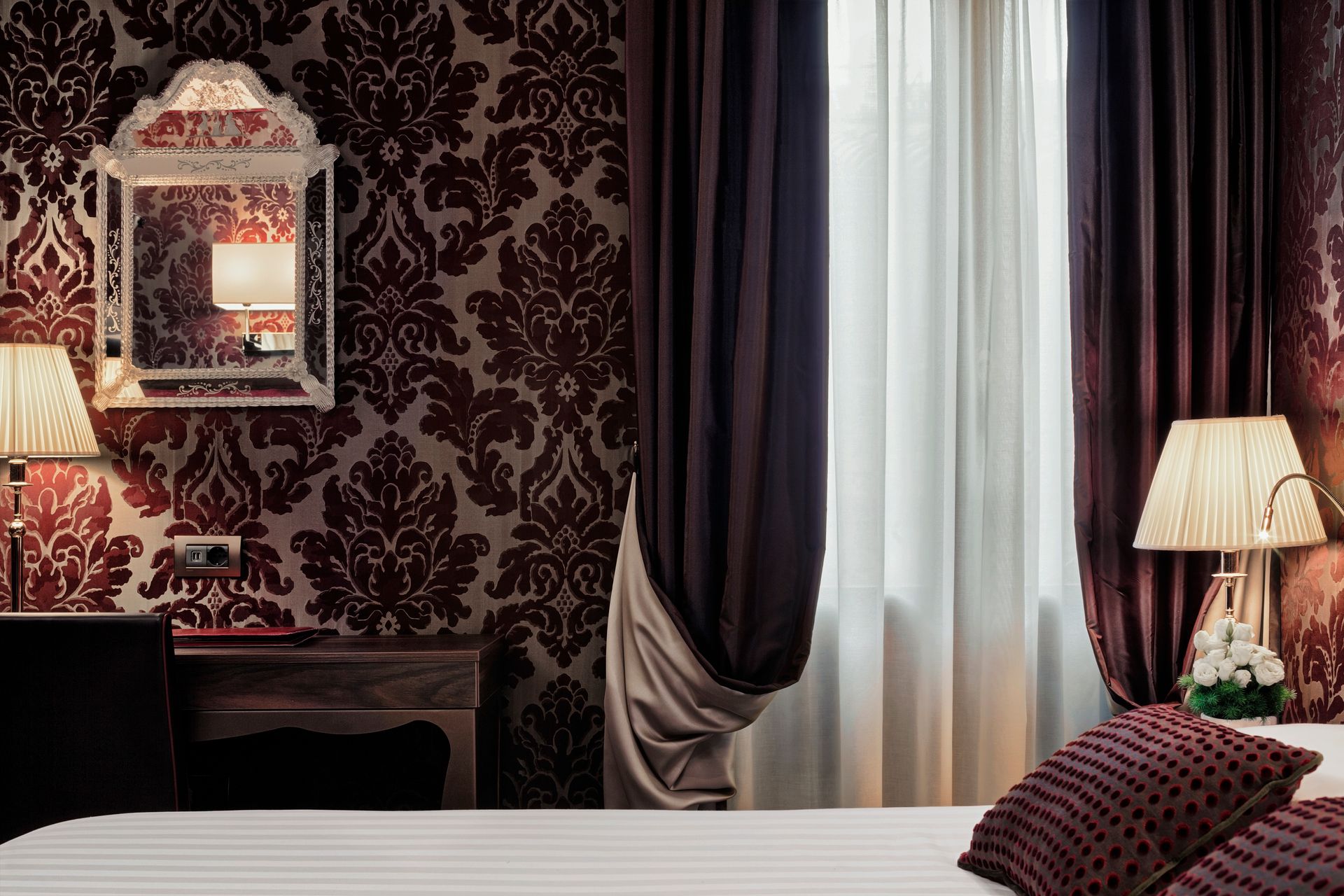 Elegante camera da letto con carta da parati scura, specchio, lampada da comodino e tende trasparenti sopra un letto bianco.