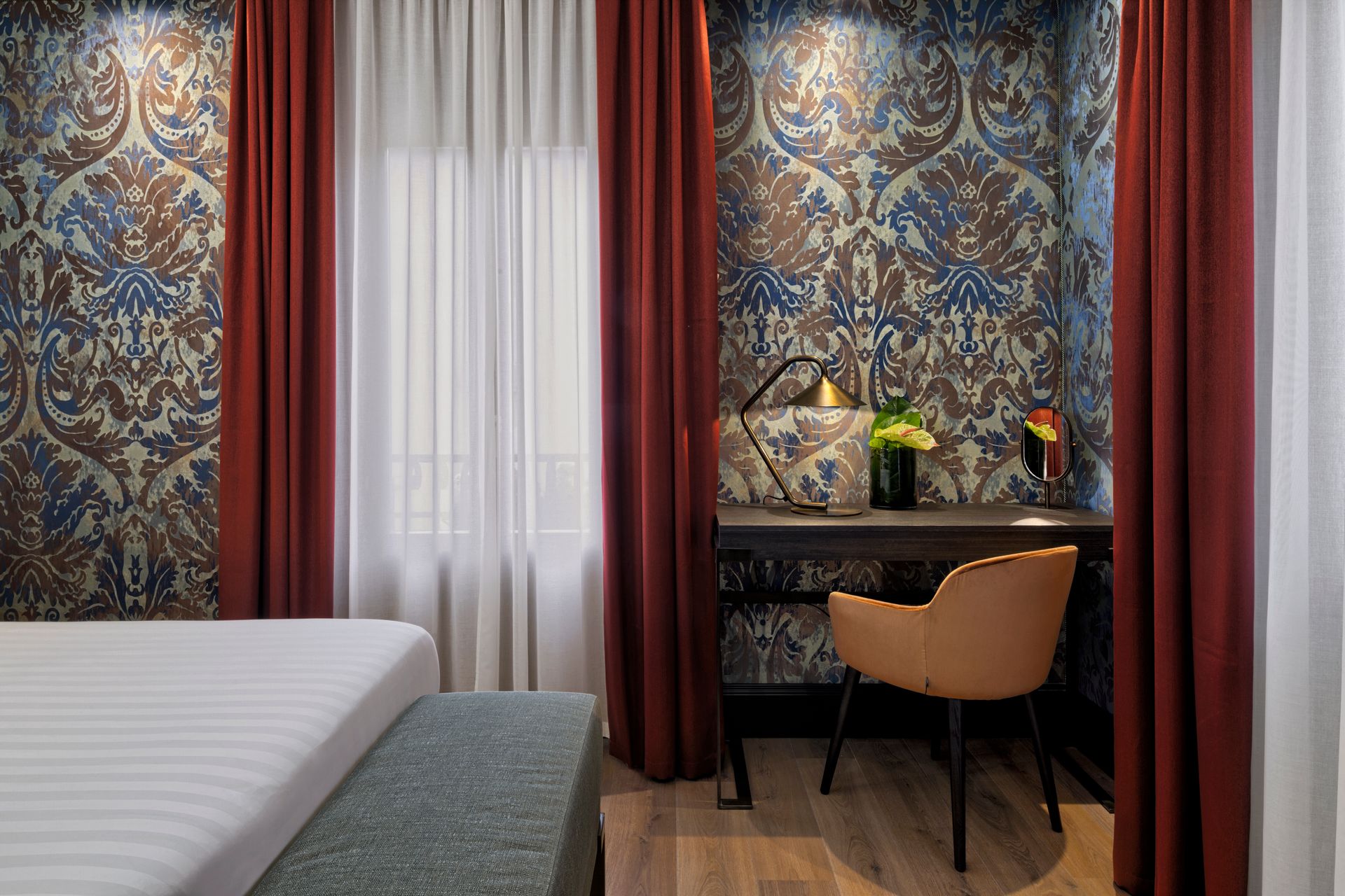 Accogliente angolo camera da letto con carta da parati a motivi, letto, scrivania, sedia e tende trasparenti.
