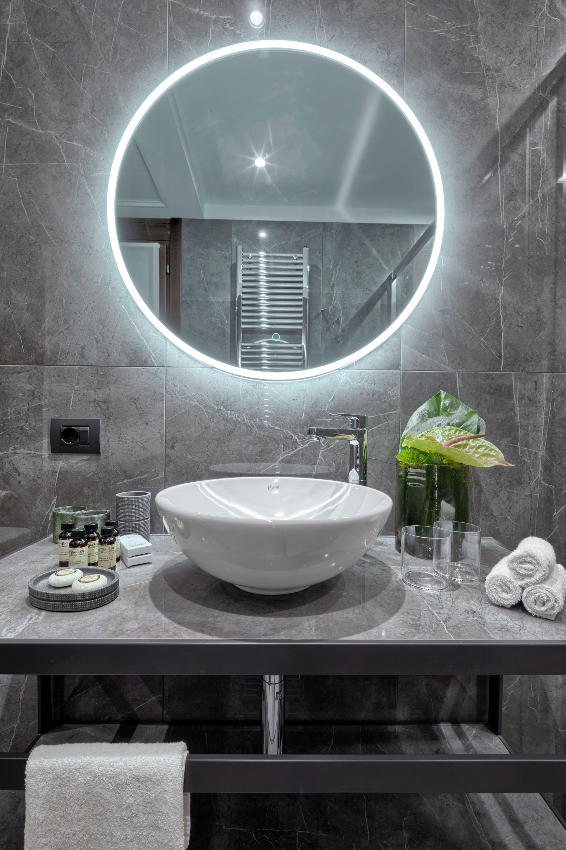 Mobile bagno moderno di colore grigio con specchio rotondo retroilluminato, lavabo bianco, fiori e asciugamani.
