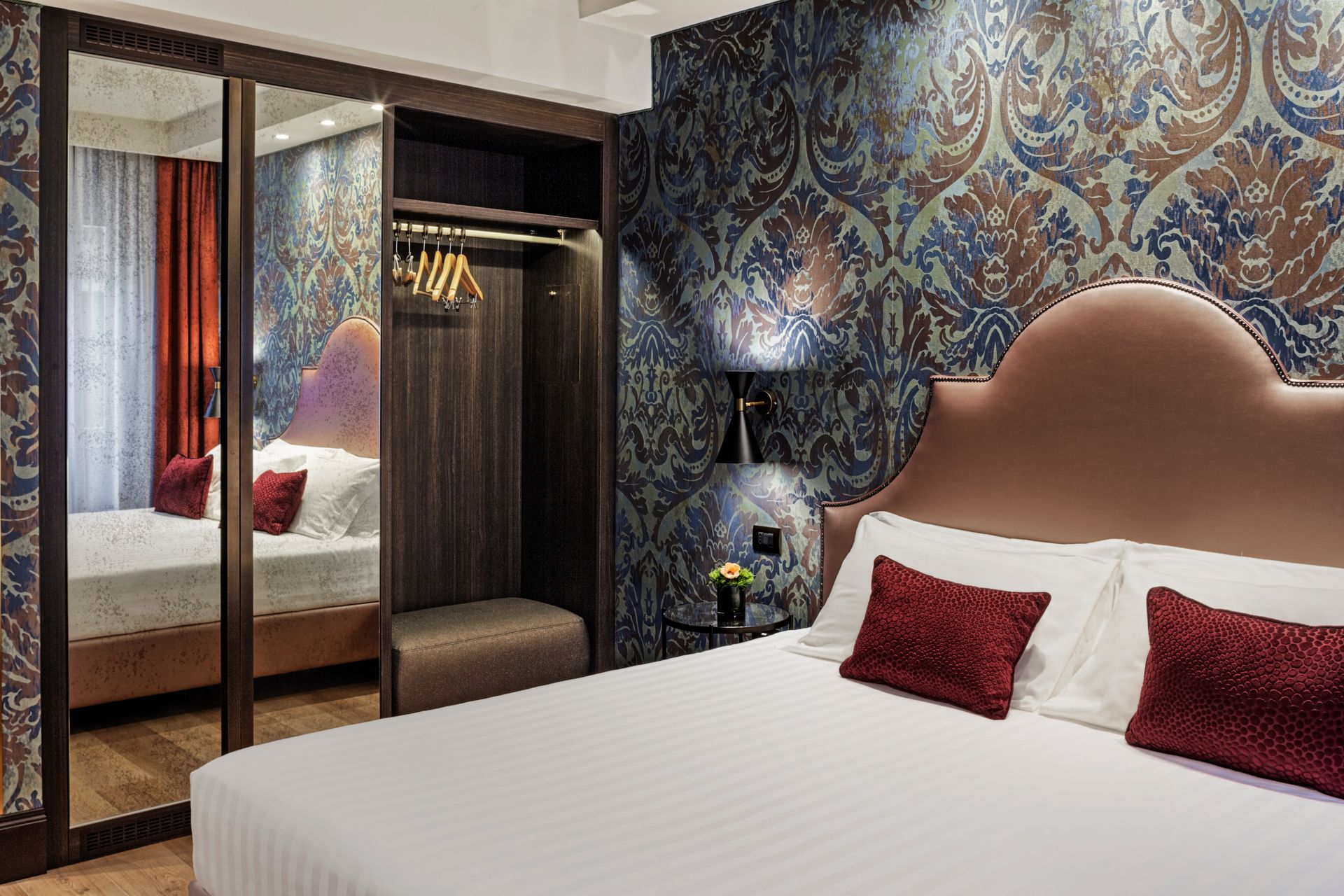 Elegante camera d'albergo con carta da parati a motivi geometrici, armadio con specchio e letto con cuscini decorativi rossi.