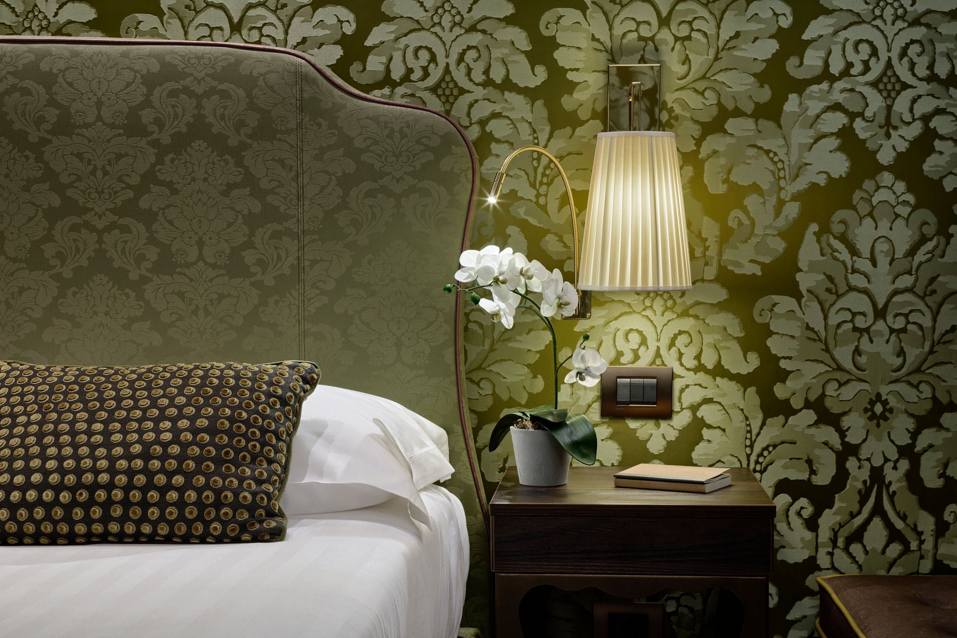 Elegante angolo di una camera d'albergo con carta da parati floreale verde, letto, lampada da comodino e orchidee bianche.