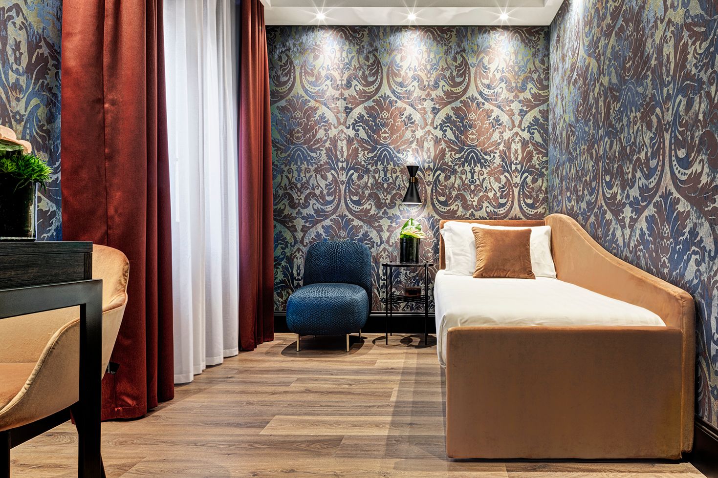 Accogliente camera da letto moderna con carta da parati a motivi, letto beige, sedia blu e tende a tutta altezza.