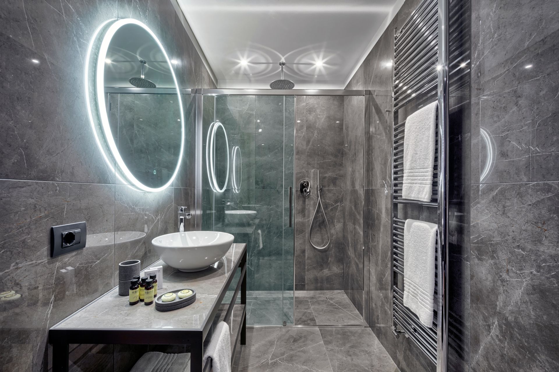 Bagno moderno di colore grigio con specchio rotondo illuminato, lavabo, doccia e portasciugamani.