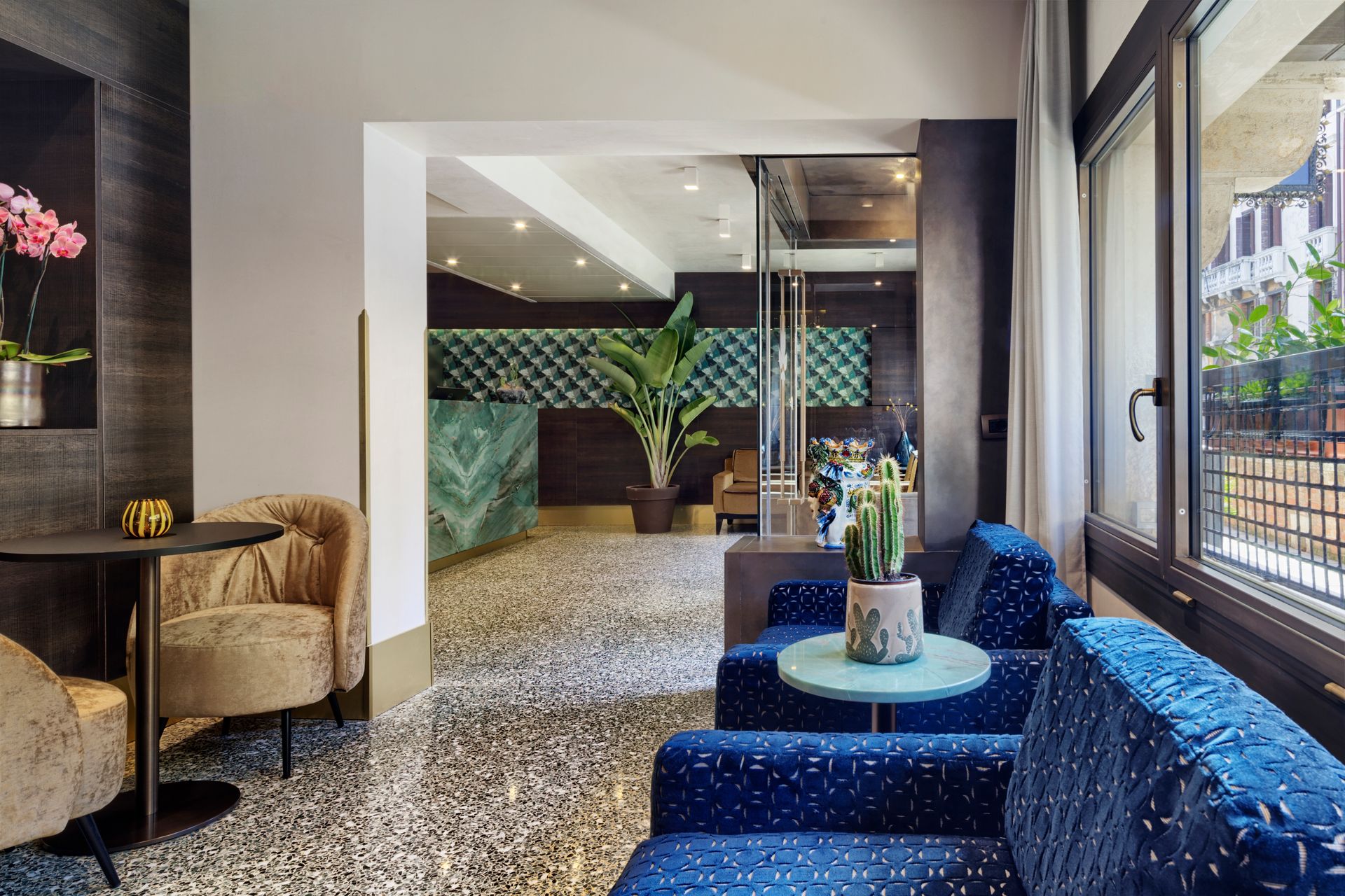 Salotto moderno con divani a fantasia blu, sedie intrecciate e pavimento in terrazzo illuminato da luci calde.