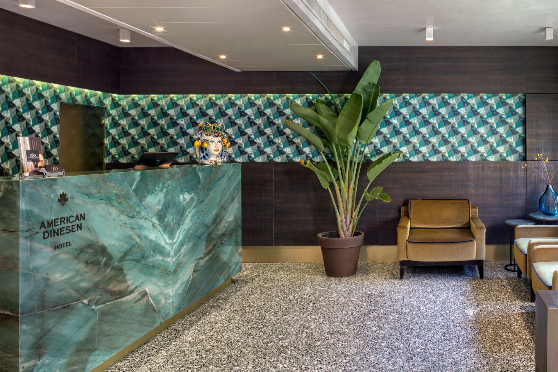 Hall dell'hotel con bancone della reception in marmo color verde acqua, pianta in vaso e area salotto su pavimento maculato.