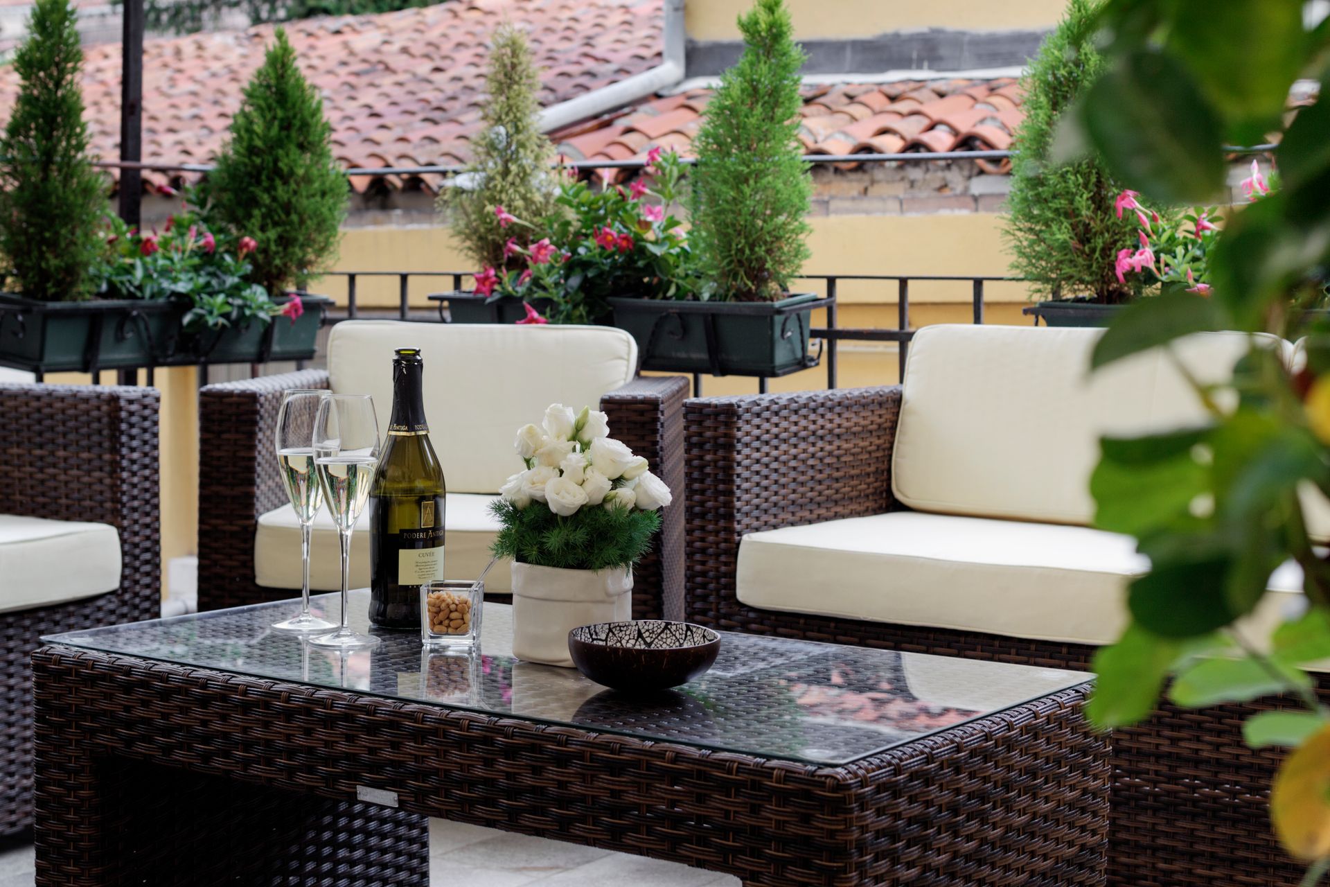 Terrasse extérieure aménagée avec canapés en osier, table basse en verre, bouteilles de vin et plantes en pot.