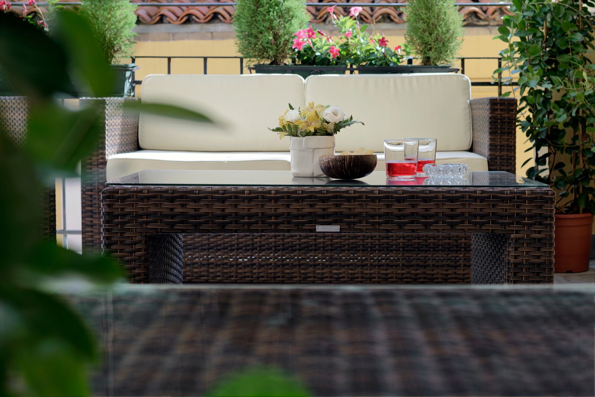Patio esterno con divano in vimini, cuscini, tavolino e piante in vaso sullo sfondo.