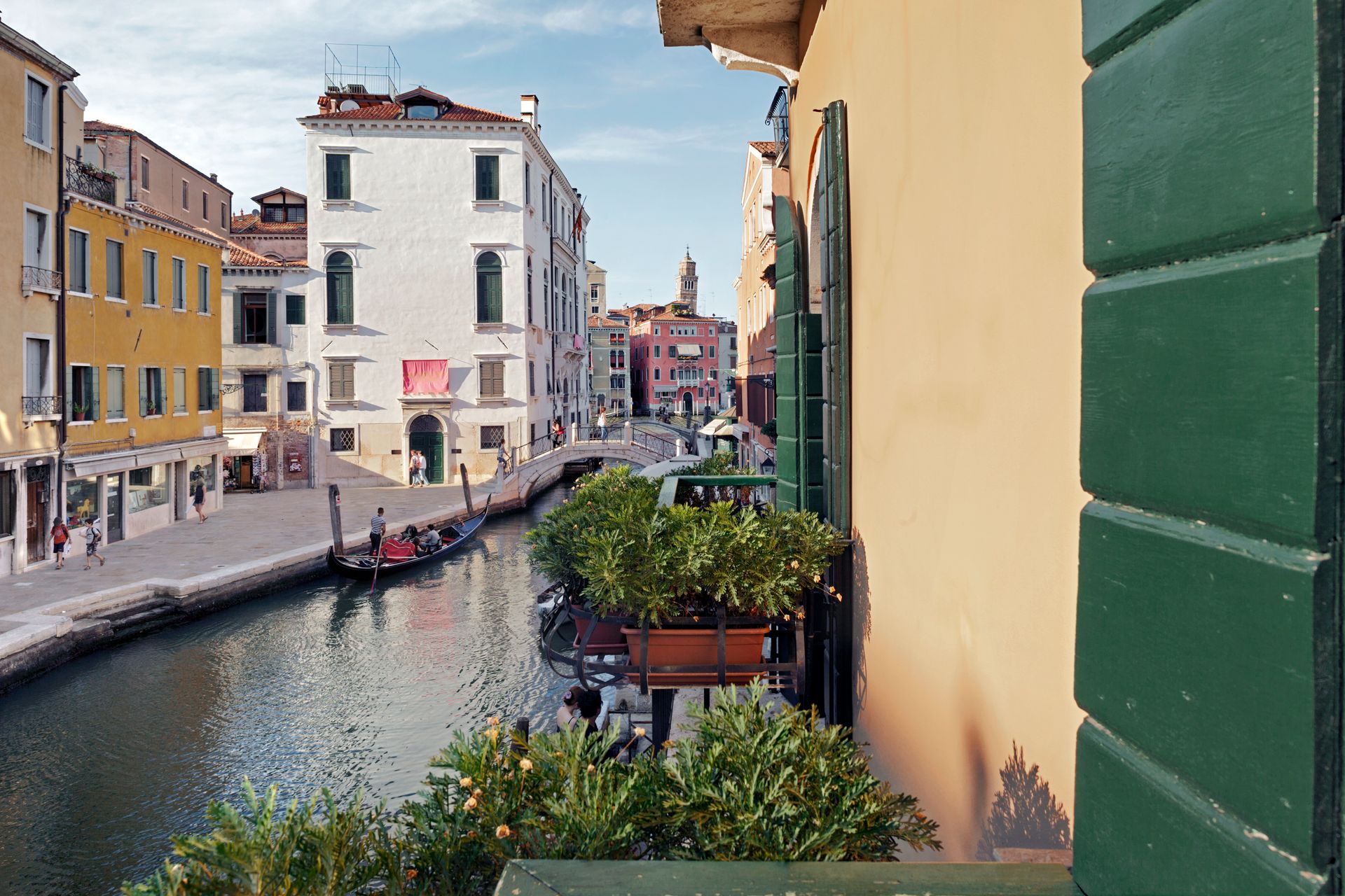 Il canale, fiancheggiato da edifici colorati, si vede da un balcone con persiane verdi e piante in vaso.
