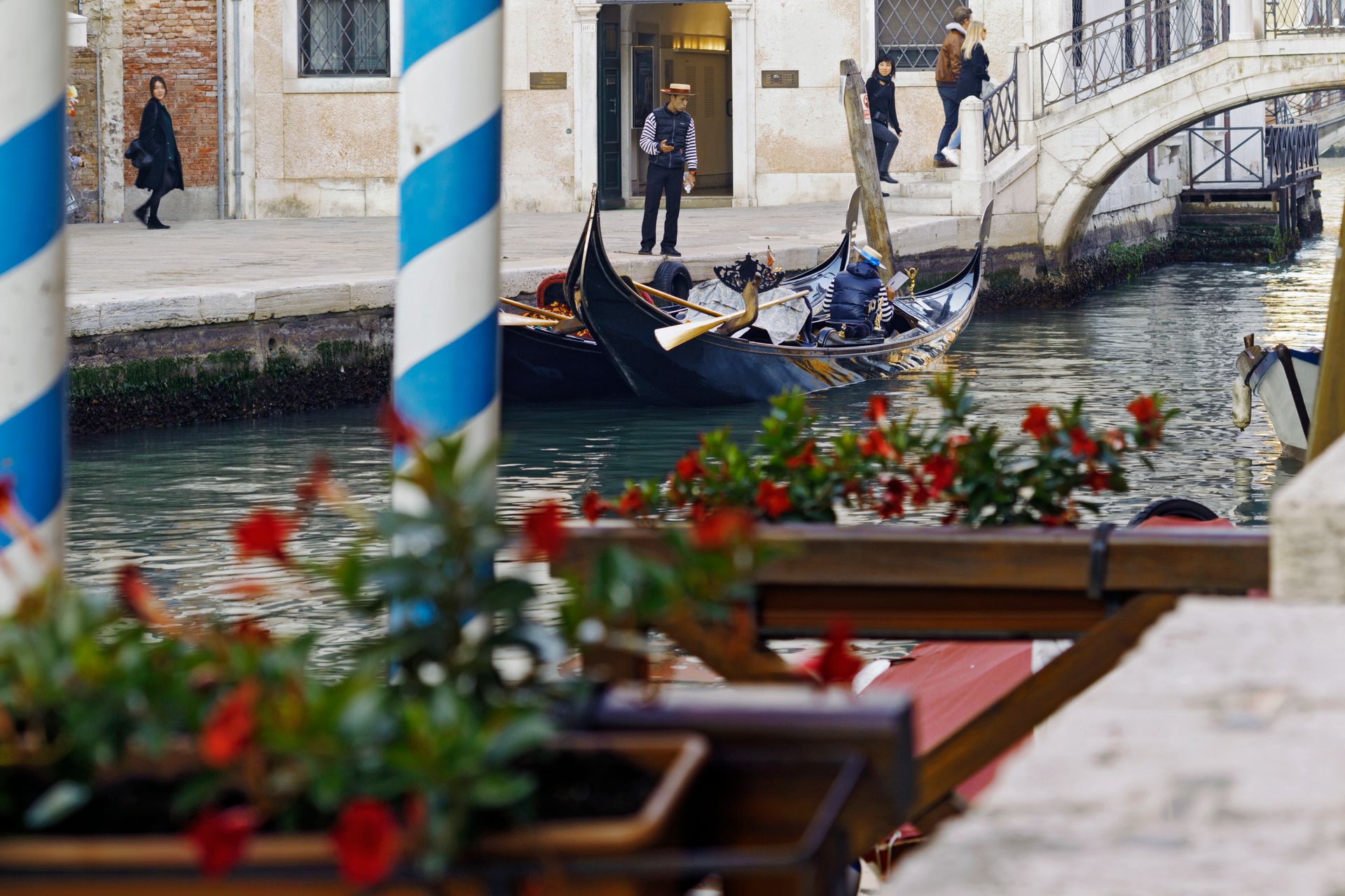 Scena di canale con gondole sotto un ponte, pali a strisce rosse e tavolini da caffè e fiori sfocati in primo piano.