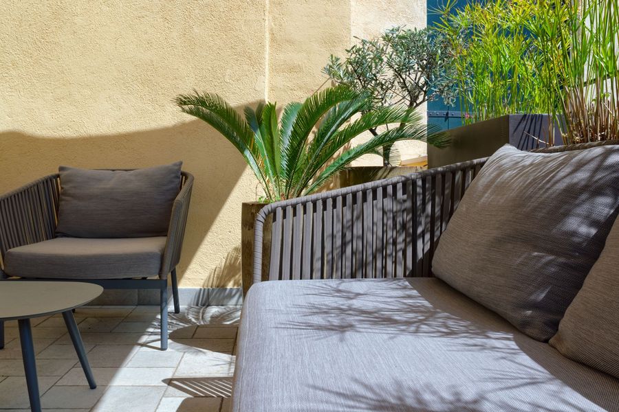 Patio esterno con sedie in vimini, cuscini color crema e un grande ombrellone in un giardino interno.